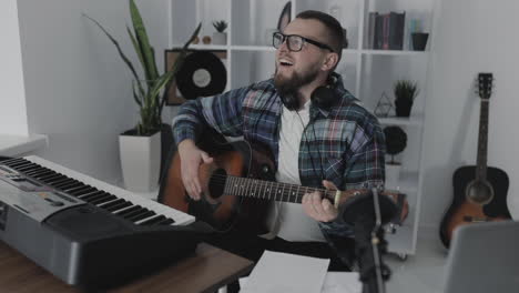 Músico-Masculino-Cantando-Y-Tocando-La-Guitarra-Junto-A-Su-Teclado-Eléctrico-En-Casa