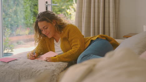 woman in cosy warm jumper lying on sofa at home lying on sofa writing greetings card or letter