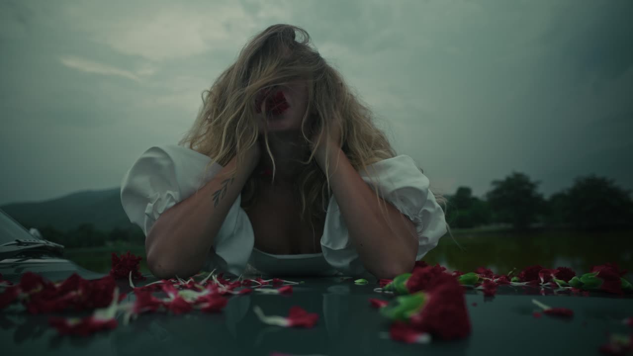 Woman in distress on a car hood amidst flowers under a gloomy sky