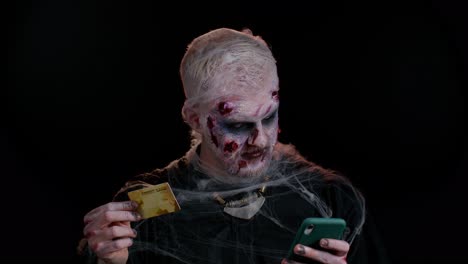 Sinister-man-Halloween-zombie-using-credit-bank-card-and-smartphone-while-purchases-online-shopping