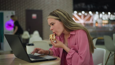 mujer de vestido rosa comiendo una hamburguesa mientras trabaja en su portátil en una mesa en un restaurante, una taza de café se coloca al lado de la computadora portátil, con personas ligeramente borrosas en el fondo mientras pasan