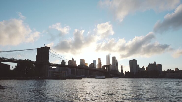 NYC: Ponte di Brooklyn e lo skyline di Downtown Manhattan da Dumbo, Brooklyn, al tramonto con nuvole che si muovono velocemente che avvolgono la guglia del One World Trade Center, e l'East River - New York City, USA.
