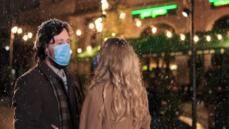 caucasian happy couple wearing facial masks talking while snowing on the street in christmas