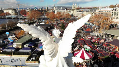 Drone-shot-of-angel-statue-in-front-of-Christmas-Market-in-Zurich,-Switzerland-during-the-day-with-the-cityscape-in-the-background