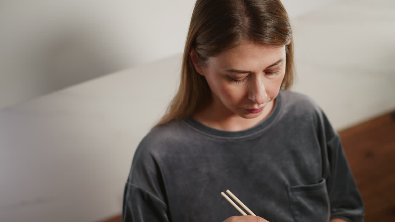 White lady in casual gray shirt holding chopsticks, eating meat with calm expression, enjoying her meal in bright modern kitchen with soft light, wooden countertop visible in background