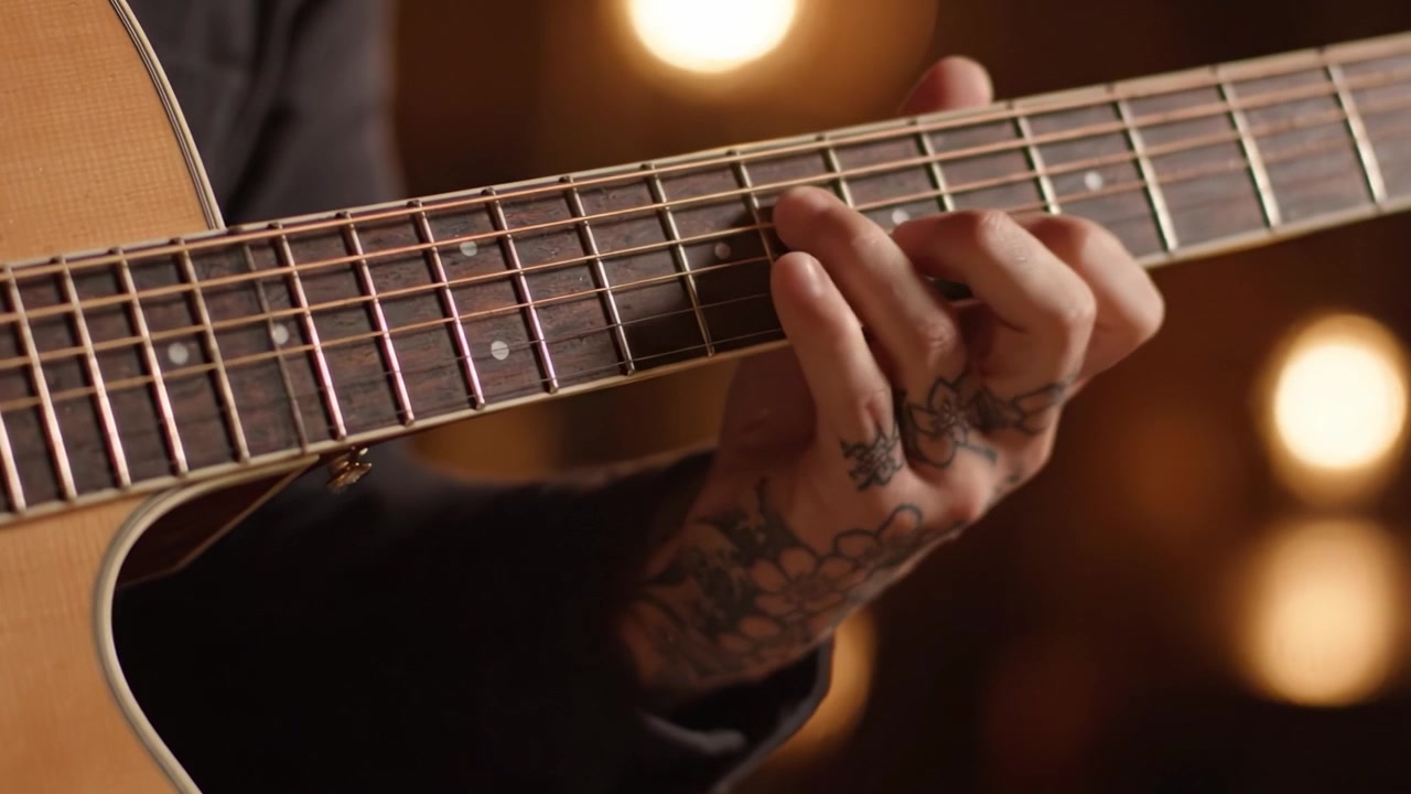 Una prospettiva ravvicinata sul suonare la chitarra: una mano tatuata che strimpella abilmente le corde sotto luci ambientali calde in un ambiente da studio musicale.
