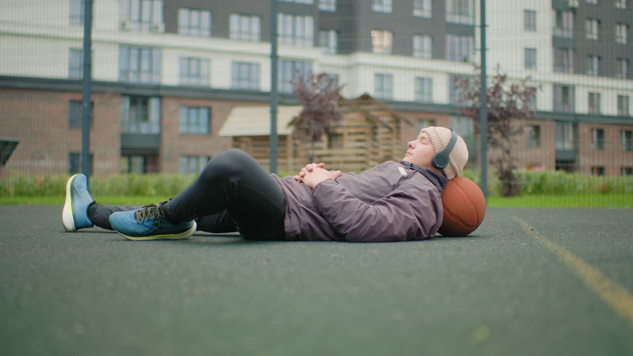Eine Person liegt auf einem Basketballfeld, hört Musik und benutzt einen Basketball als Kissen. Im Hintergrund befinden sich bewölkte Wohnhäuser. Die Person trägt einen Hoodie, Turnschuhe und Kopfhörer, ist nachdenklich und urban.