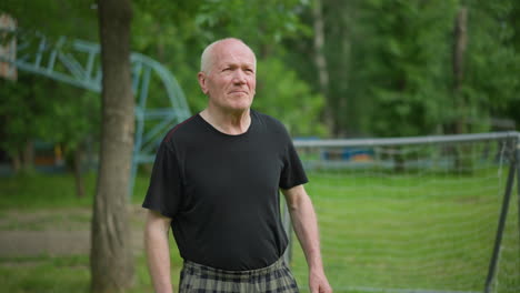 an elderly man throws a soccer ball with determination and focus, showcasing his energy and intent, with a blurred background with a goalpost, trees, and a building