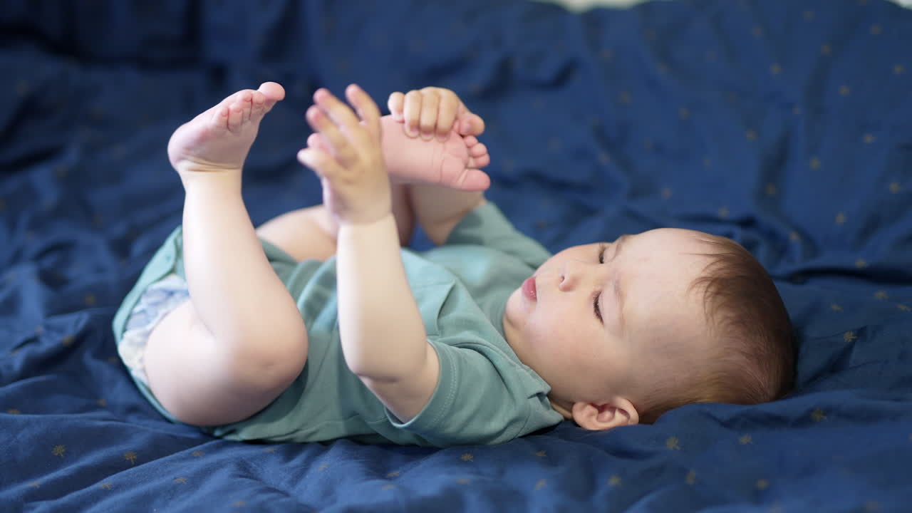 Cute lovely kid being focused on his feet. Toddler boy playing with his feet while lying on the blue bedclothes. Side view.