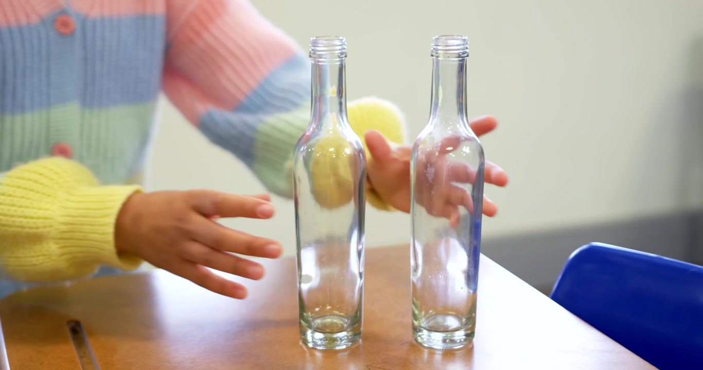African American teen in pastel top seeing two bottles, recycling into red bin at classroom desk
