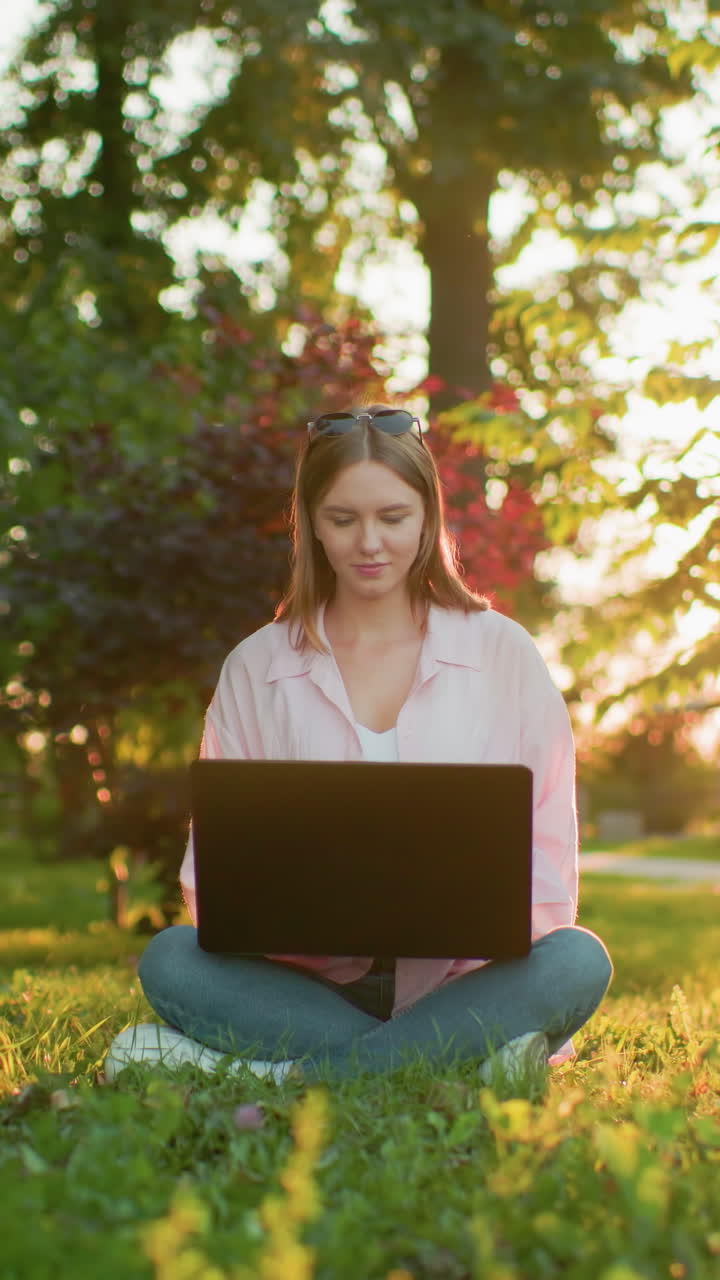 Freelancer works on laptop in park, seated cross-legged with sun creating a golden halo effect, lush greenery and filtered sunlight in background enhance serene setting