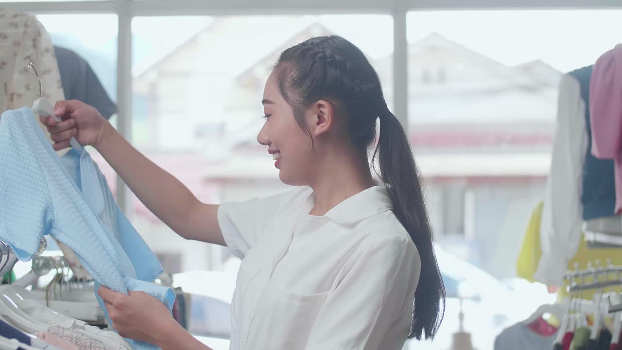 부티크 에서 쇼핑 하는 동안 옷 을 입으려고 하는 미소 짓는 젊은 아시아 여자