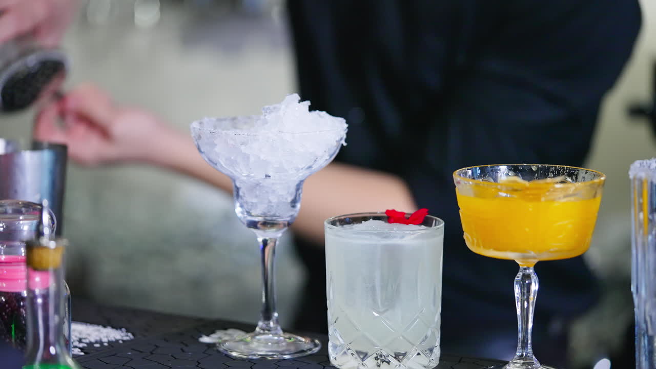 Beautiful diverse glasses filled with ice and drinks. Barman in black shirt preparing cocktails at backdrop in blur.