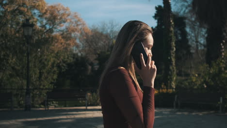 Side-view-shot-of-young-woman-in-red-top-talking-on-smartphone-walking-through-park-by-the-lake.