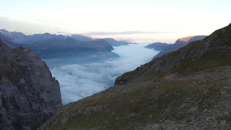 Drone-shot-flying-sideways-to-the-left,-showing-rocks,-stones-and-pebbles-on-the-slope-of-the-mountain,-getting-steeper-into-the-cliff-that-goes-into-the-massive-valley-with-fog,-mist-and-clouds