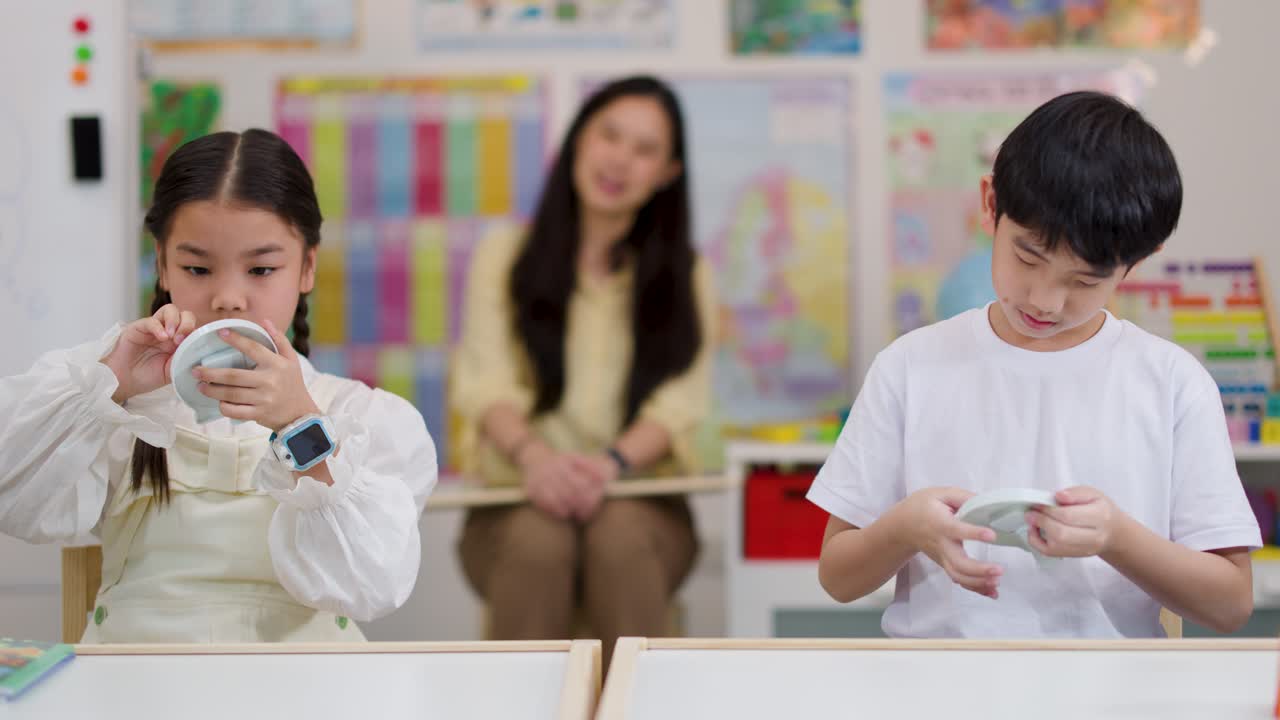 Two children adjust toy clocks in a bright classroom, supervised by a teacher, steady camera