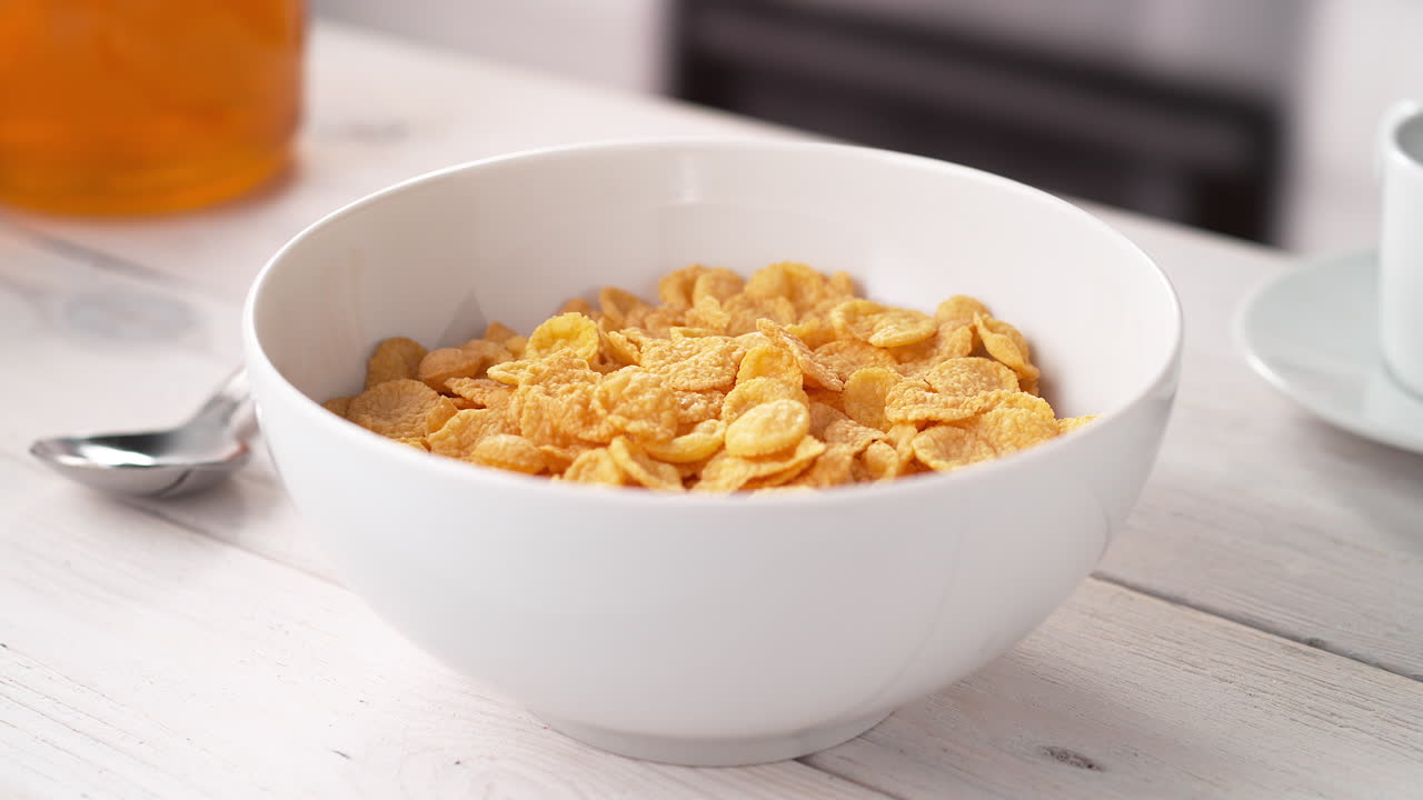 preparar el desayuno: verter leche en un cuenco de copos de maíz dorados de cereal colocados en una mesa de la cocina, cámara lenta