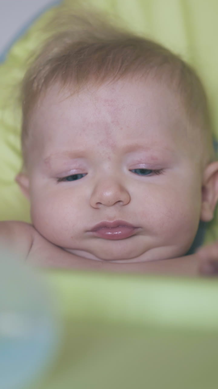 funny worried upset baby boy sits in soft green highchair with plastic bottle of water on table at home close view