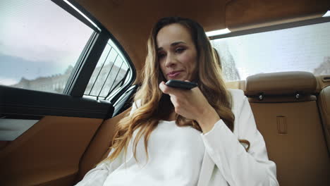 Smiling-business-lady-recording-voice-message-on-phone-in-salon-of-business-car.