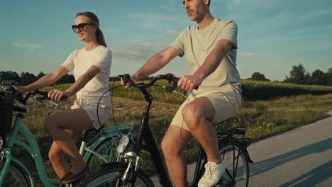 Vista-Lateral-De-Una-Juguetona-Pareja-Caucásica-Divirtiéndose-Mientras-Anda-En-Bicicleta-Al-Atardecer-En-La-Carretera-Del-Pueblo.