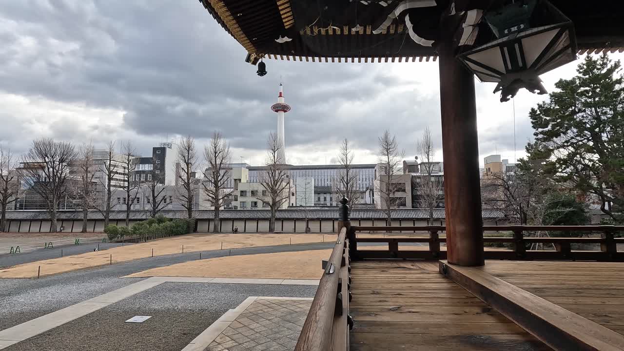 View of Kyoto Tower seen from the wooden veranda of Higashi Hongan-ji Temple, with cloudy sky and bare winter trees.