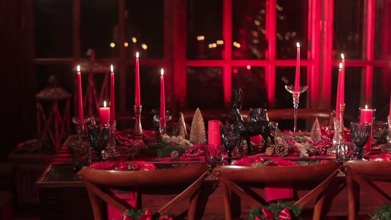 Beautifully decorated table for festive Christmas dinner. Burning candles, pine cones, figurines and luxurious crockery in the room lit in red color.