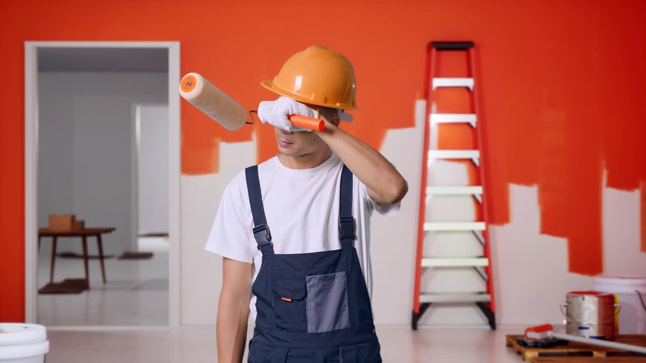 pintor hombre asiático con casco de seguridad limpiando el sudor y estando cansado mientras está de pie con la pared de pintura en la habitación