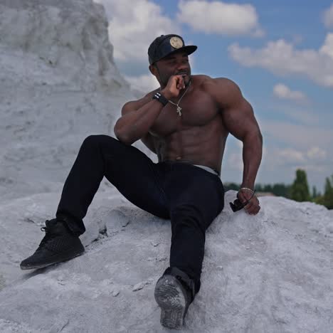 thoughtful black skinned guy sitting on white stone. shirtless african american man with muscular body rests outdoors. sportsman on hill in summer