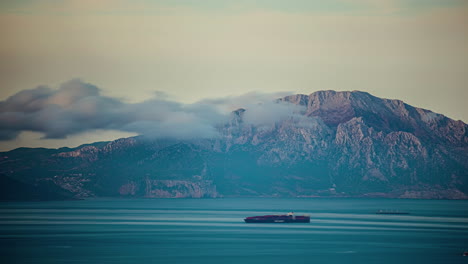 boats and clouds pass over a blue sea with mountains in the background