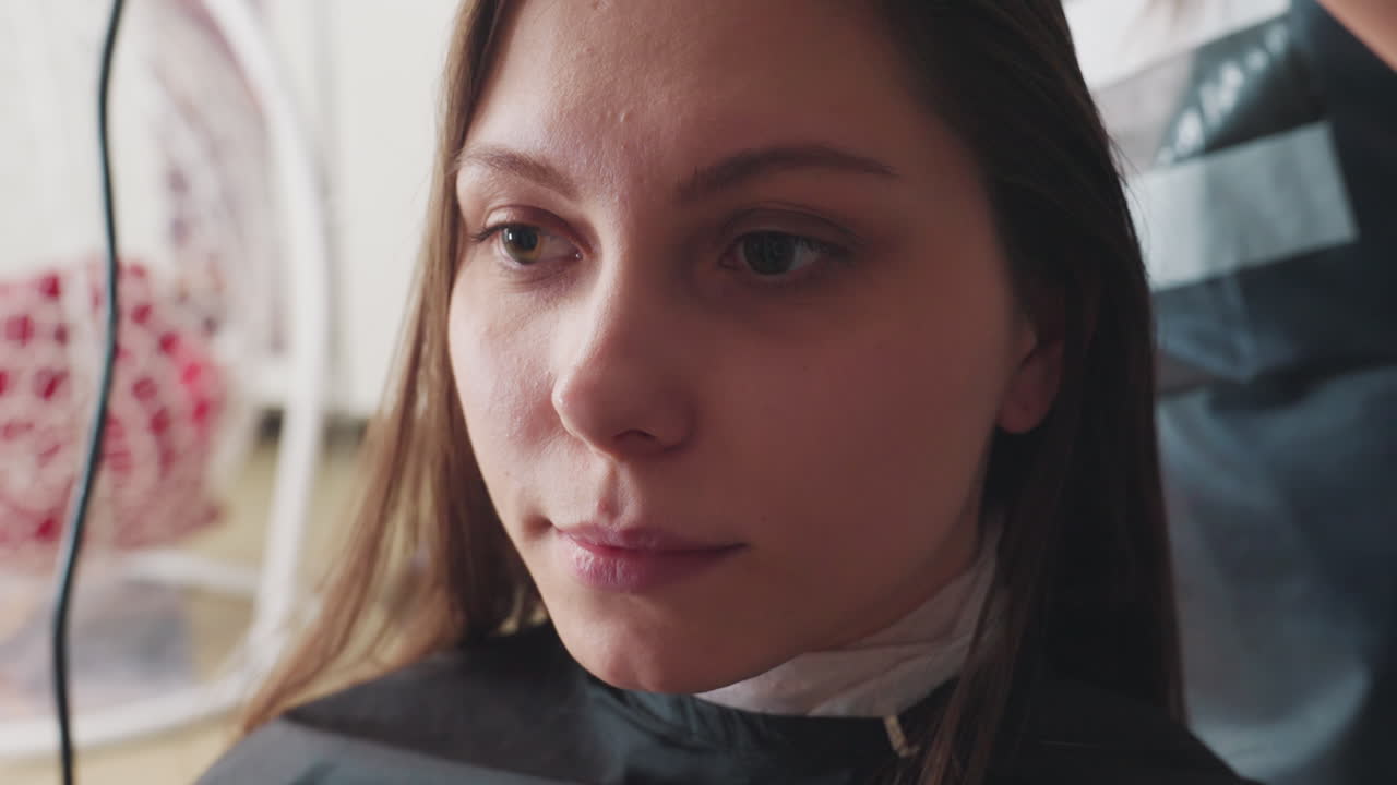 Cliente esperando con tranquilidad, persona sentada con una capa, individuo con capa de peluquería mostrando una expresión facial natural, retrato de una persona relajándose en una silla de peluquería con capa durante el proceso de peinado