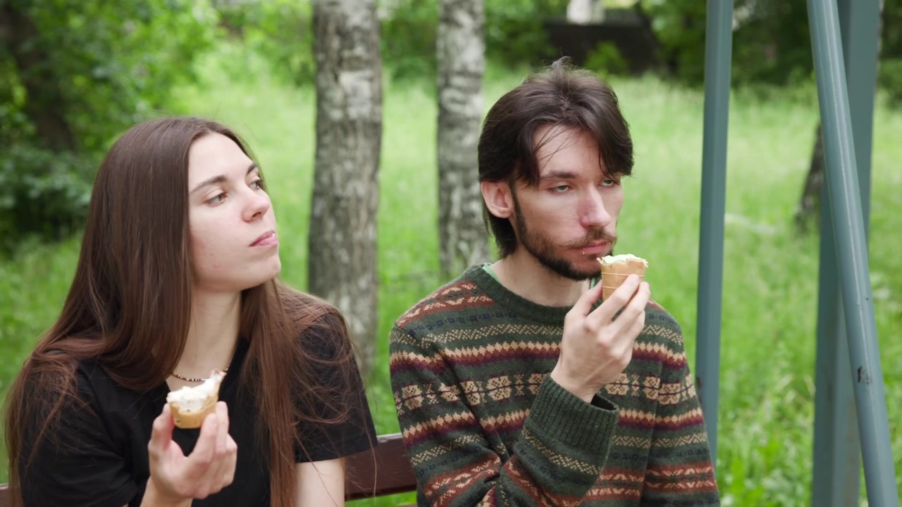 Two young adults sit side by side on wooden bench in grassy birch grove, each holding small wafer ice cream cone, eating and turning faces in soft daylight with relaxed outdoor mood