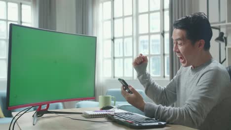 asian man in long sleeved t-shirt with green screen desktop computer looking at mobile phone and being happy successful in work while working at home.