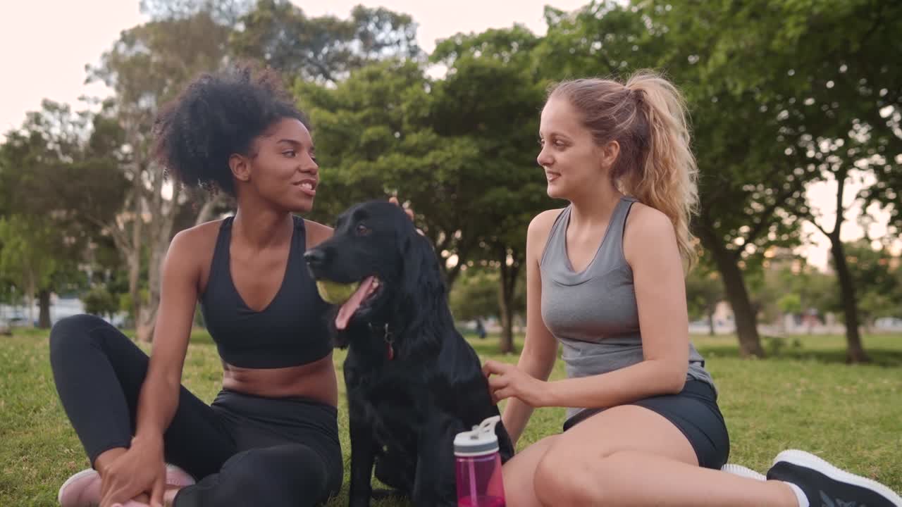 dos mujeres jóvenes de fitness en ropa deportiva relajándose junto con su perro mascota después de la sesión de entrenamiento en el parque