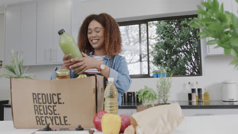 Mujer-Birracial-Feliz-Desempacando-Comestibles-De-Una-Caja-Reciclada-En-La-Cocina,-Espacio-Para-Copiar,-Cámara-Lenta