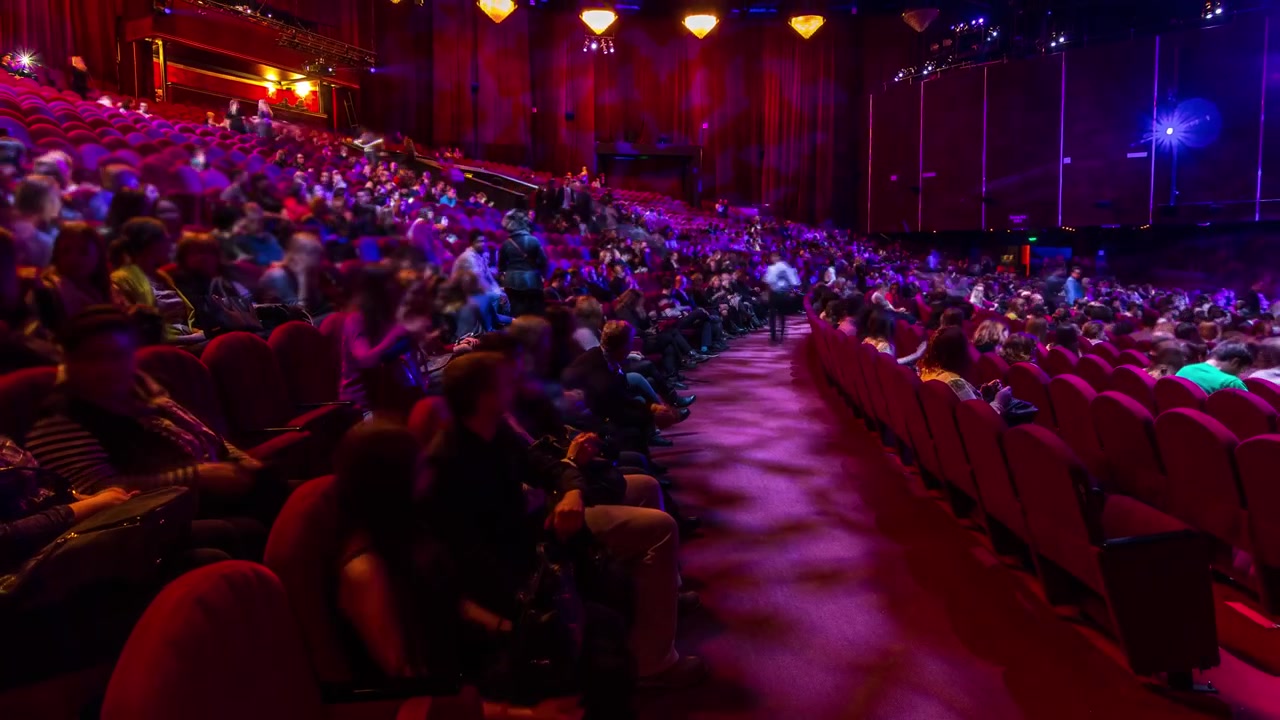 Spectators gather in the auditorium and watch the show in theatre timelapse. Large hall with red armchairs seats. Viewers filling places until turn off the light. Panoramic view from left side