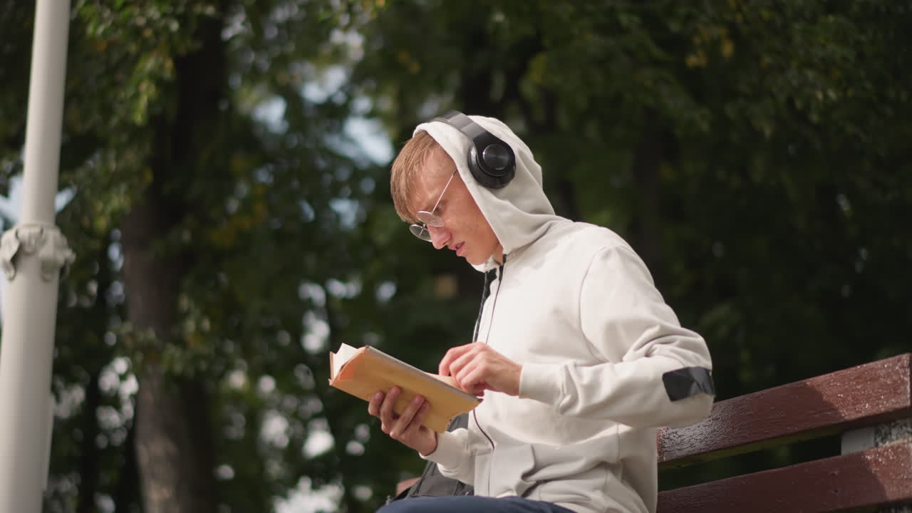 Seorang pelajar kulit putih asyik membaca di bangku, close-up frame memperlihatkan ekspresi fokus, headphone di kepala, hoodie, buku terbuka, cahaya belang-belang menembus pepohonan, konsentrasi tenang, peran bergeser