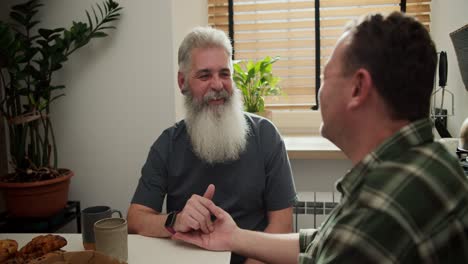 Ein-Glücklicher-Mann-Mit-Grauem-Haar-Und-üppigem-Grauem-Bart-Im-Fortgeschrittenen-Alter-In-Einem-Grauen-T-Shirt-Kommuniziert-Mit-Seinem-Brünetten-Freund-In-Einem-Karierten-Grünen-Hemd-Und-Streichelt-Seine-Hand-Während-Eines-Gesprächs-Mit-Einem-LGBT-Paar,-Das-Beim-Mittagessen-In-Der-Wohnung-Am-Tisch-Sitzt