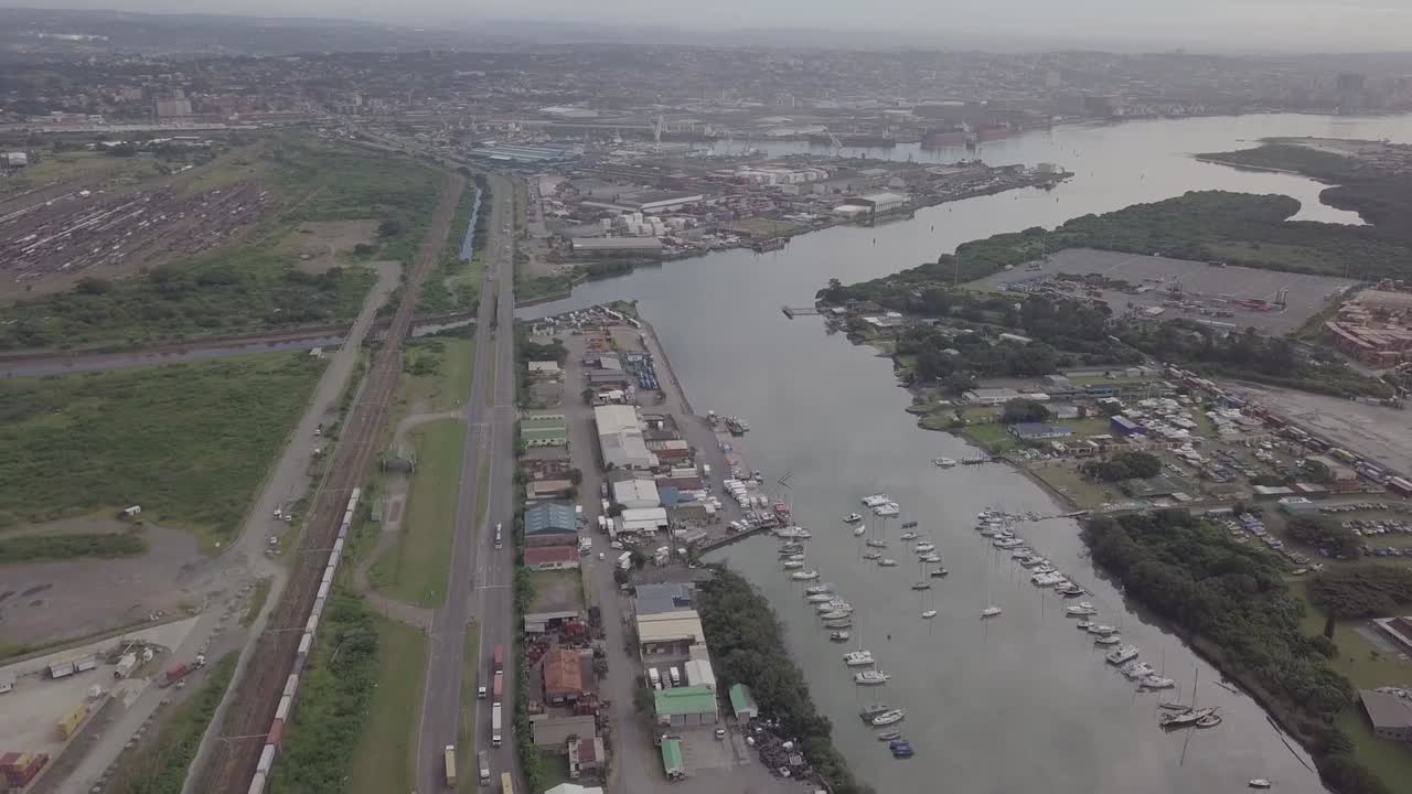 Aerial footage of Durban harbour with yachts and traffic consisting of trucks and moving trains over a highway with cargo container depot in view.