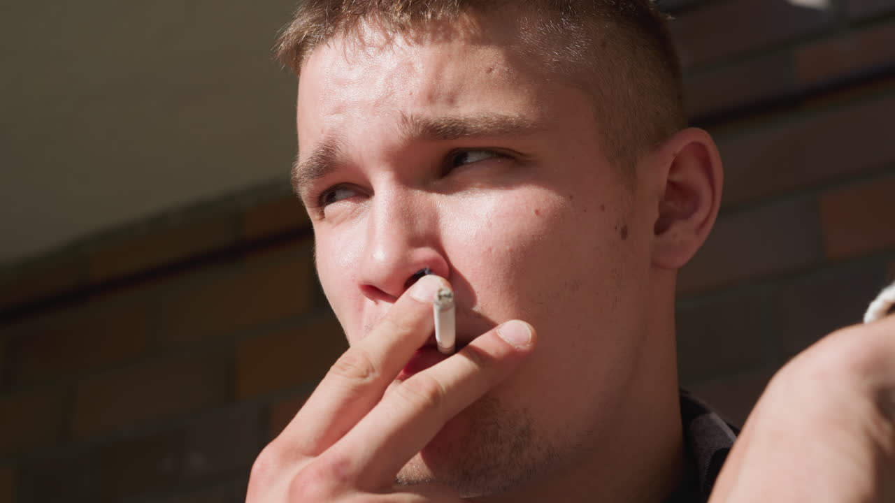 Close up man in black white checkered shirt smoking cigarette sunlight glints off face as he lifts filter to lips holding fresh lit cigarette with blurred brick background