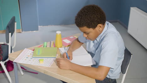 american boy sitting at desk and writing in notebook during class at school