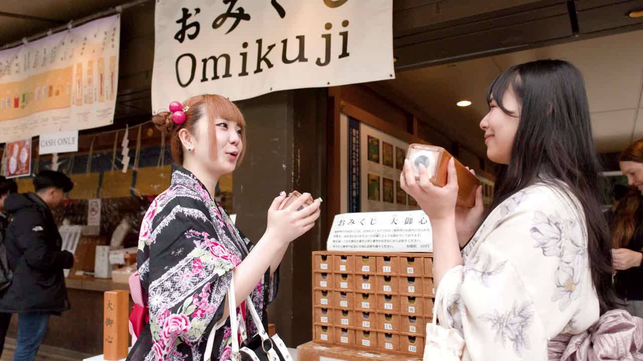 To kvinder iført traditionelle kimonoer ryster heldigkasser i træ ved en japansk helligdom i Tokyo