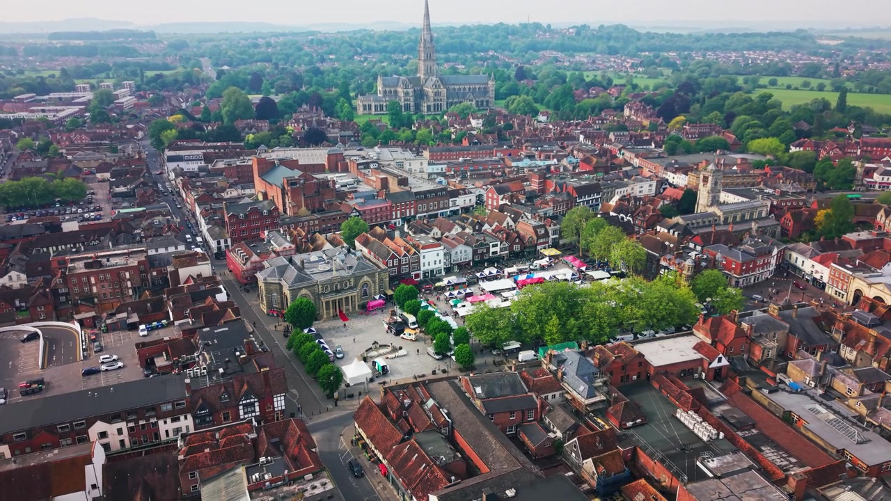 Drone footage looking across Salisbury Market Square with market stalls crowds the Guildhall St Thomas Church historic rooftops trees and Salisbury Cathedral in the distance.