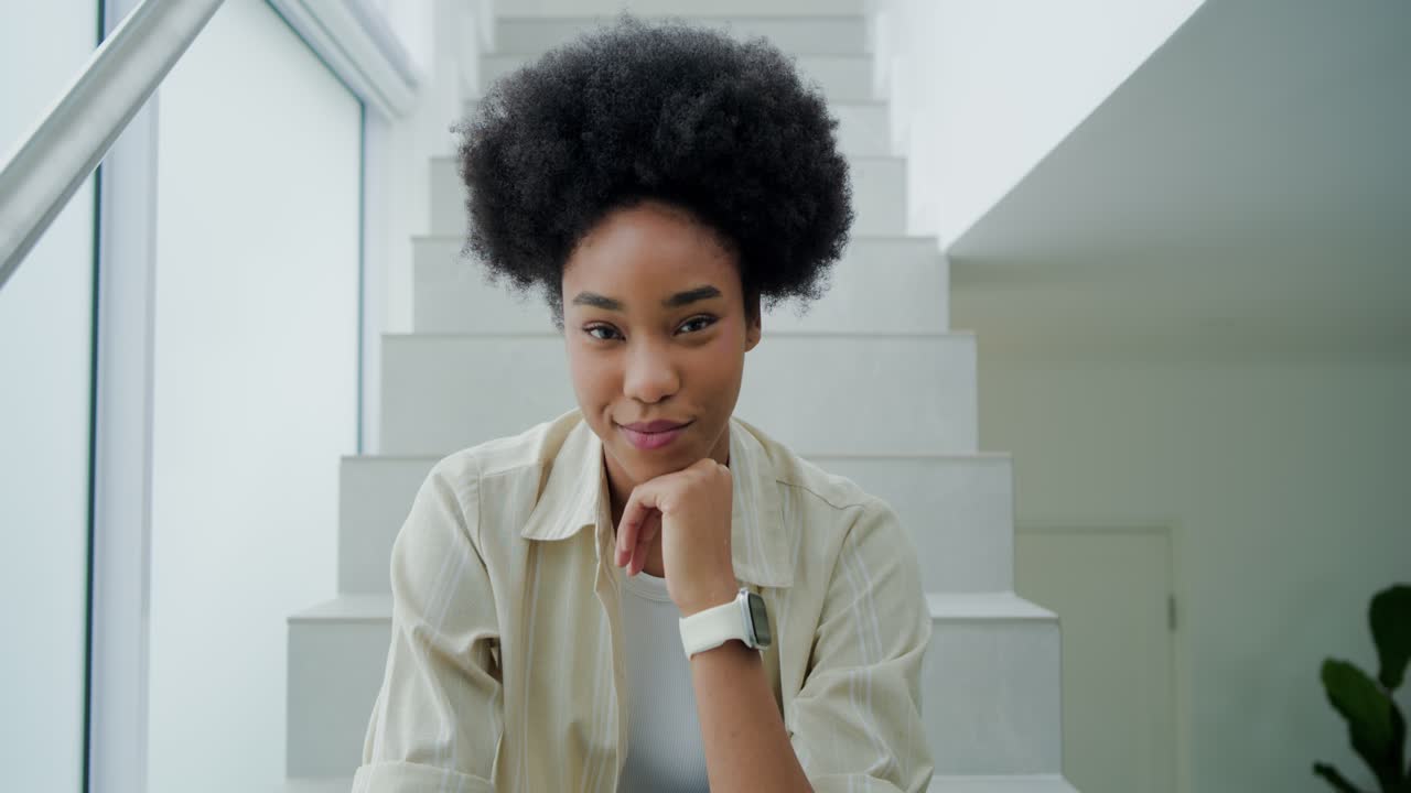 African American woman in twenties sitting on stairs by window wearing beige shirt and smartwatch