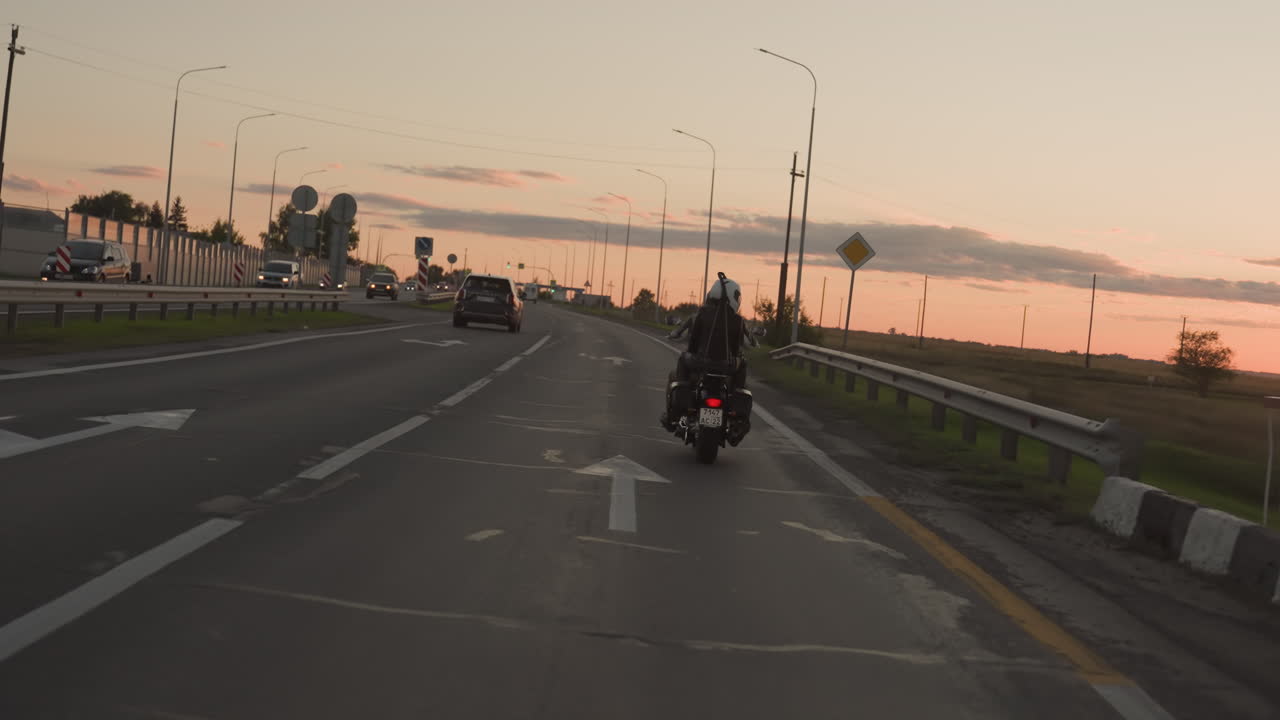 Motorcycle with rider and passenger travels along highway during sunset, following road signs and vehicles under evening sky, symbolizing freedom, motion, travel, and adventure lifestyle on open road