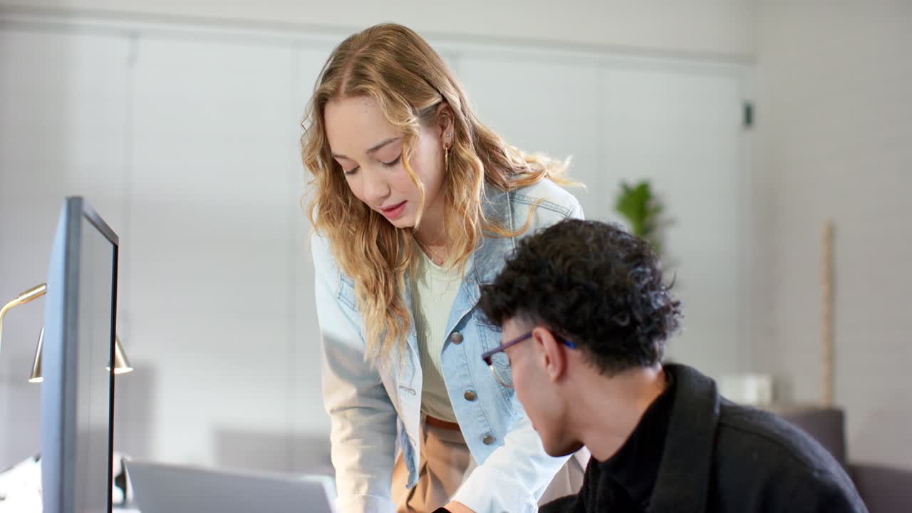 Colegas de trabalho diversos em uma mesa: uma mulher questiona e, em seguida, digita no monitor do laptop, orientando um homem que está inclinado.