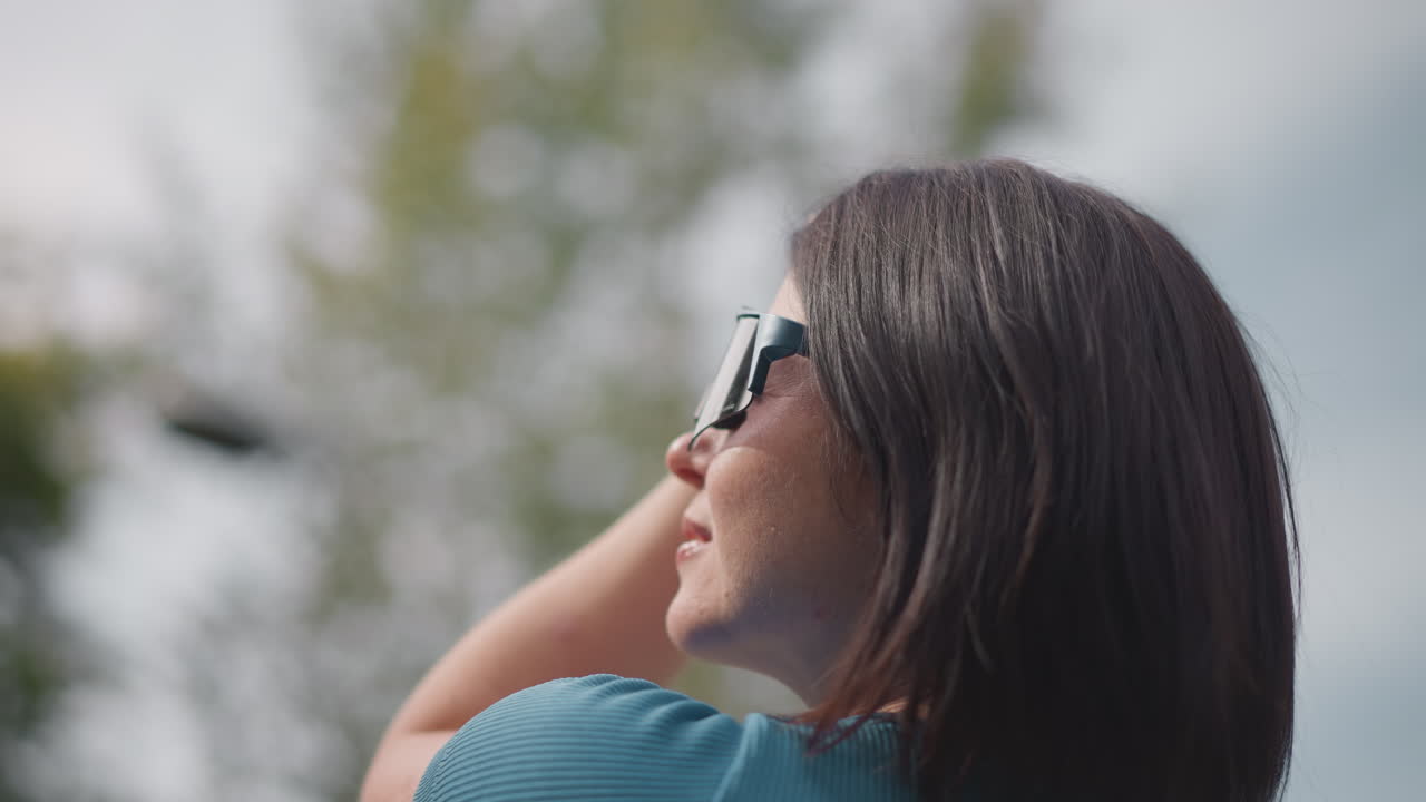 Donna di spalle con casco, che regola la cinghia e alza gli occhiali da sole in un parco tranquillo, con la luce soffusa del sole che filtra tra gli alberi; visibili un caschetto scuro e una maglietta color verde acqua, primi piani di testa e spalle.