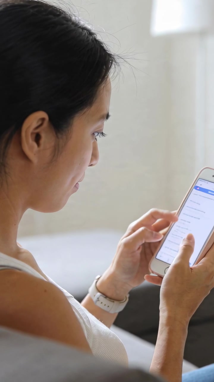 Vertical video: Tapping thumbnails, woman in sleeveless top browsing list on couch with smartphone