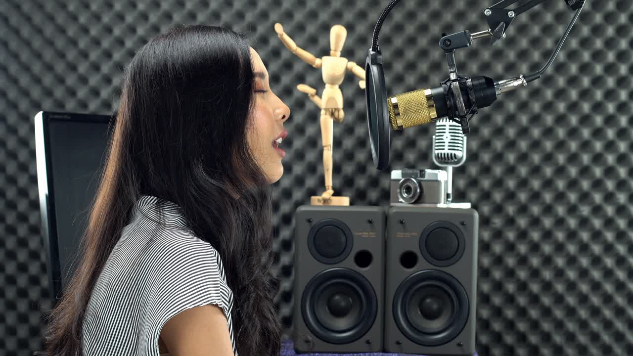 Giovane donna asiatica con lunghi capelli neri raccolti canta una canzone a gran voce con una voce potente, davanti a un microfono a condensatore sospeso e attrezzatura