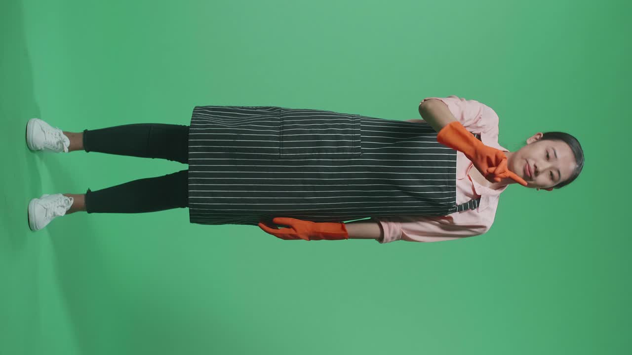 Helkropsbillede af en uenig asiatisk kvindelig husholderske med forklæde, der vifter med fingeren, mens hun står i et green screen-studie