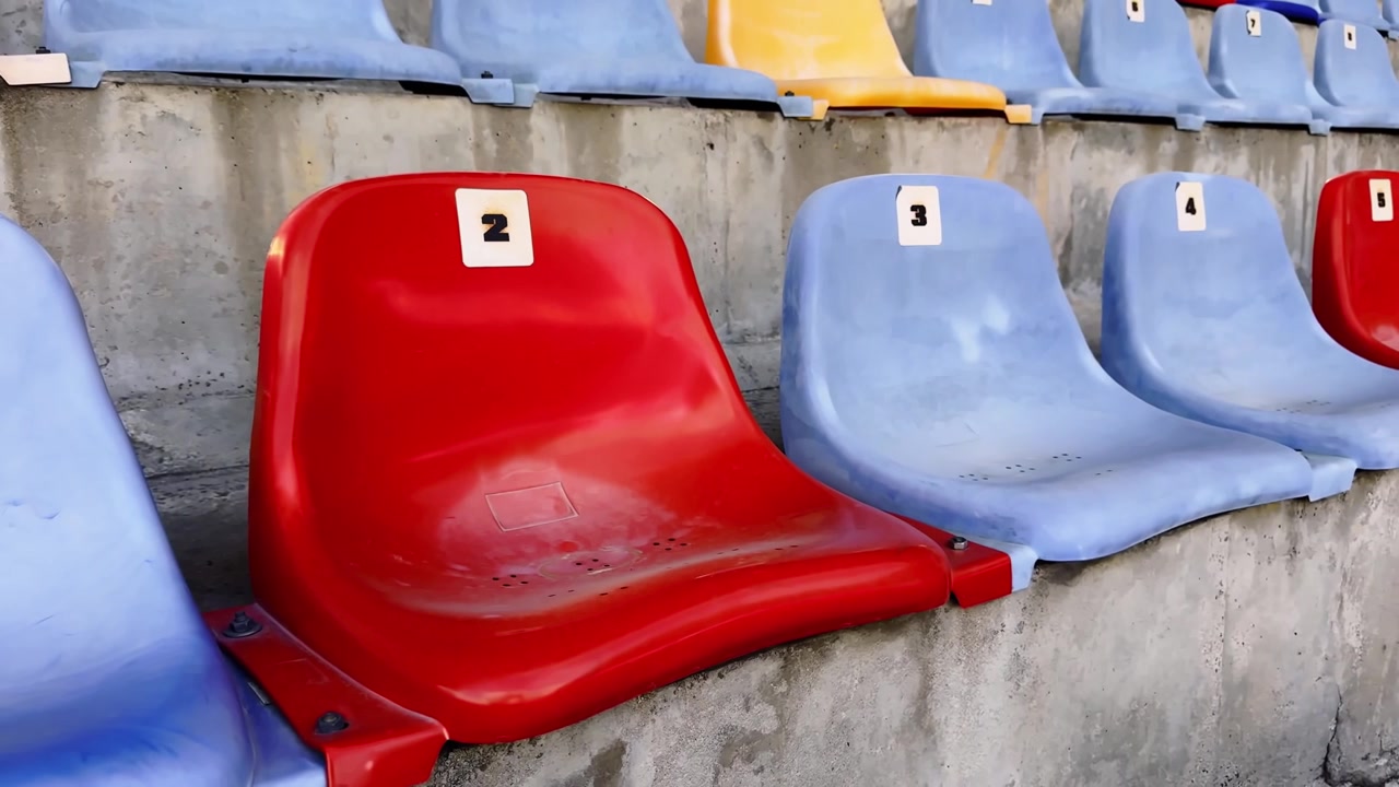 Close-up of empty stadium seats with red and blue plastic chairs, numbered labels and concrete rows in a sports venue.
